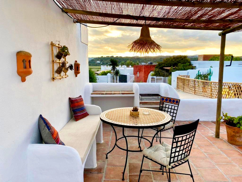 A tranquil terrace space is presented, featuring a circular table surrounded by black and white chairs. A white sofa adorned with colorful cushions is nestled against the wall. The area is shaded by a woven cover, with soft lighting and lush greenery visible in the background.