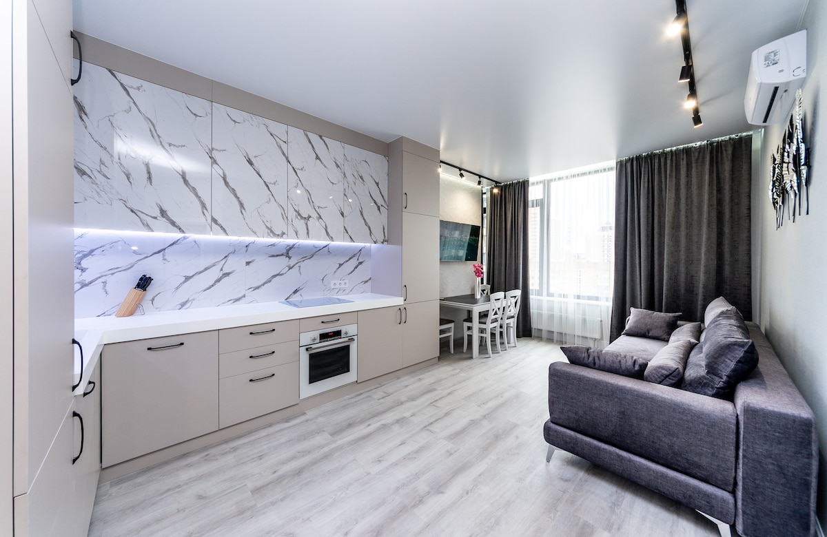 A modern kitchen area features a light marble backsplash with integrated lighting, surrounded by beige cabinetry. A cozy sectional sofa sits adjacent to a dining area with a white table and chairs. Large windows are dressed with dark curtains, allowing natural light to fill the space.