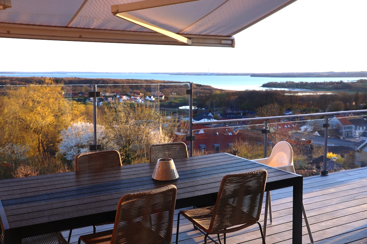 A dining table is positioned on a spacious terrace, offering panoramic views of the surrounding landscape and water. Natural light enhances the tranquil setting, while the nearby railing adds a touch of security to the outdoor space.