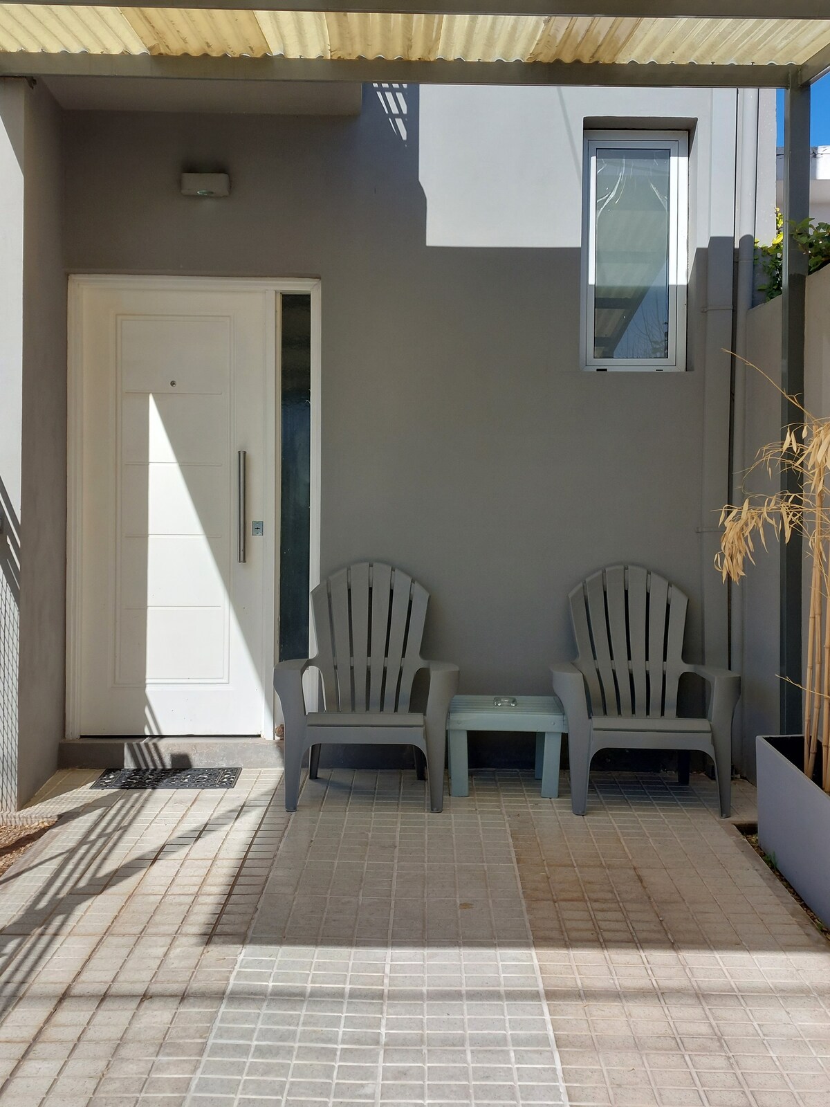 A covered entrance leads to an exterior patio area, featuring two gray Adirondack chairs and a small table. Natural light filters through a nearby window, illuminating the tiled flooring and surrounding gray walls.