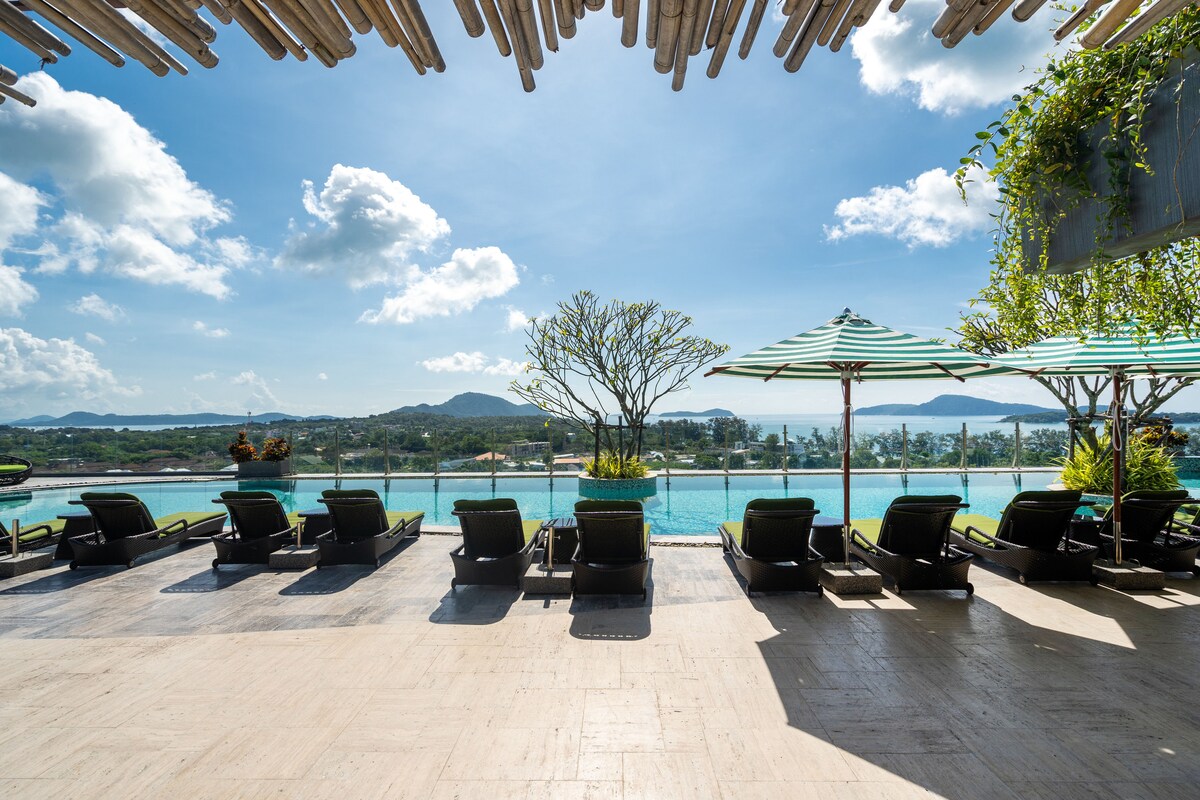 An infinity pool overlooks the sea, framed by lounge chairs arranged for relaxation. Umbrellas provide shade, while potted plants add greenery. The sky is bright with clouds, and hills are visible in the distance, creating a serene backdrop for the rooftop area.
