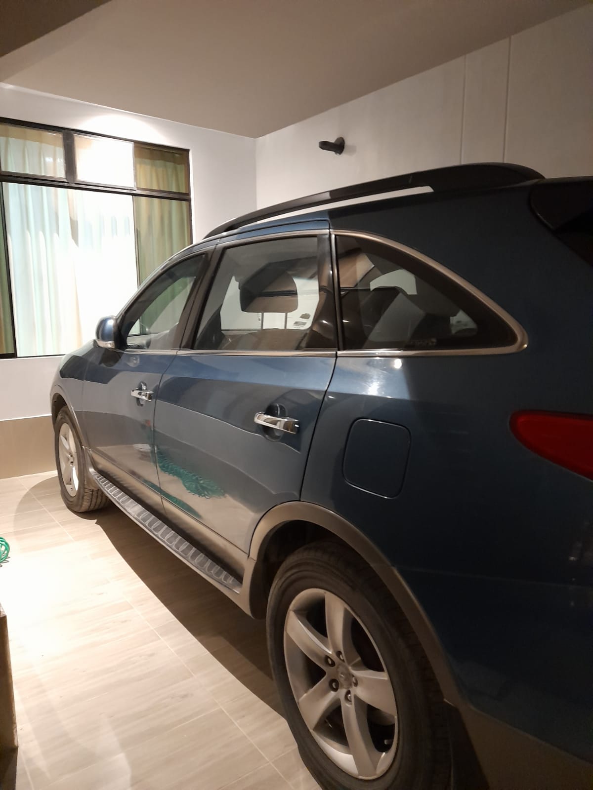 A spacious garage area is shown, featuring a parked blue SUV. Light filters in through a large window, illuminating the neutral-toned walls and tiled floor. The vehicle is positioned on the right side of the frame, with ample space surrounding it.