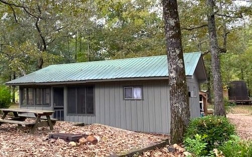 The cabin exterior is surrounded by trees, featuring a green metal roof and gray siding. A screened porch is visible on one side, and a picnic table is placed nearby, with a firepit in front among fallen leaves.