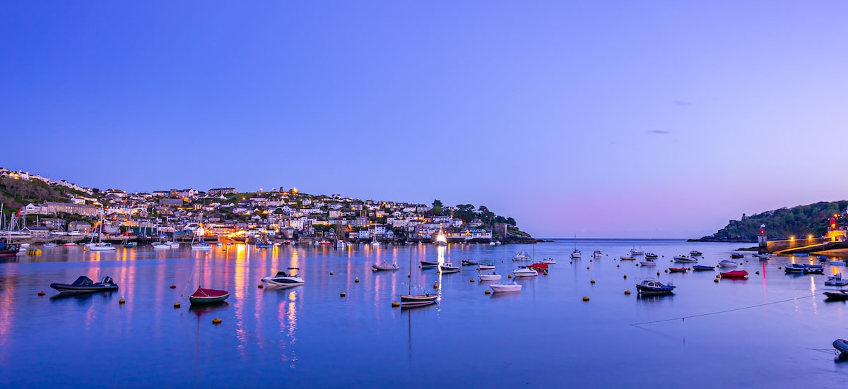 A tranquil harbor scene is presented during twilight, showcasing calm waters reflecting lights from the nearby village. Various boats are anchored, and the backdrop features hillside buildings with subtle illumination against a fading blue sky.