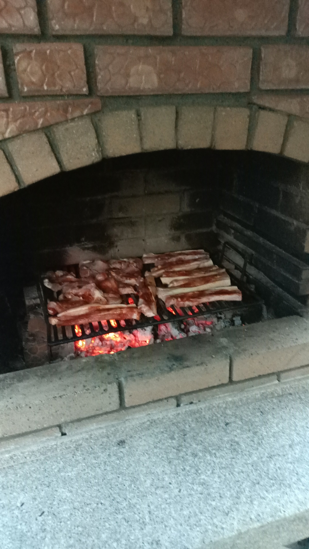 An outdoor barbecue grill is shown, featuring slabs of meat cooking over glowing red coals. The grill is constructed of stone bricks, providing a rustic appearance. The warmth of the fire is visible, enhancing the inviting setting for outdoor dining.