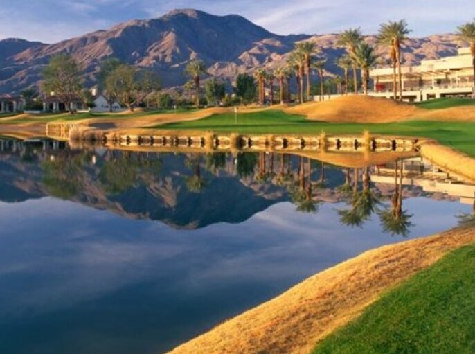 The image captures a serene golf course setting, featuring a calm water reflection of surrounding palm trees and mountains. The grassy landscape is punctuated by a gentle slope, showcasing a blend of natural and manicured elements in a tranquil atmosphere.