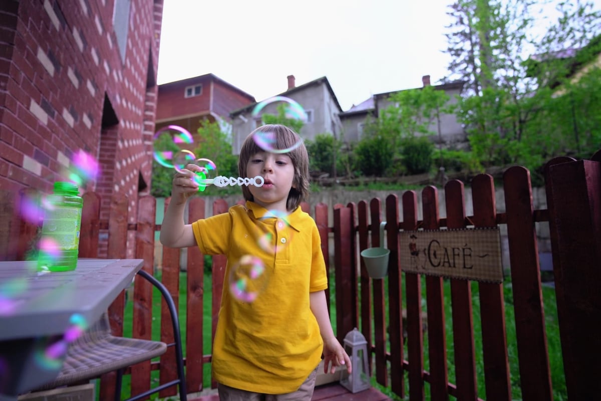 A child in a yellow shirt is blowing bubbles on a terrace, creating a playful scene. A wooden fence is visible in the foreground, while greenery and houses are seen in the background under a cloudy sky.