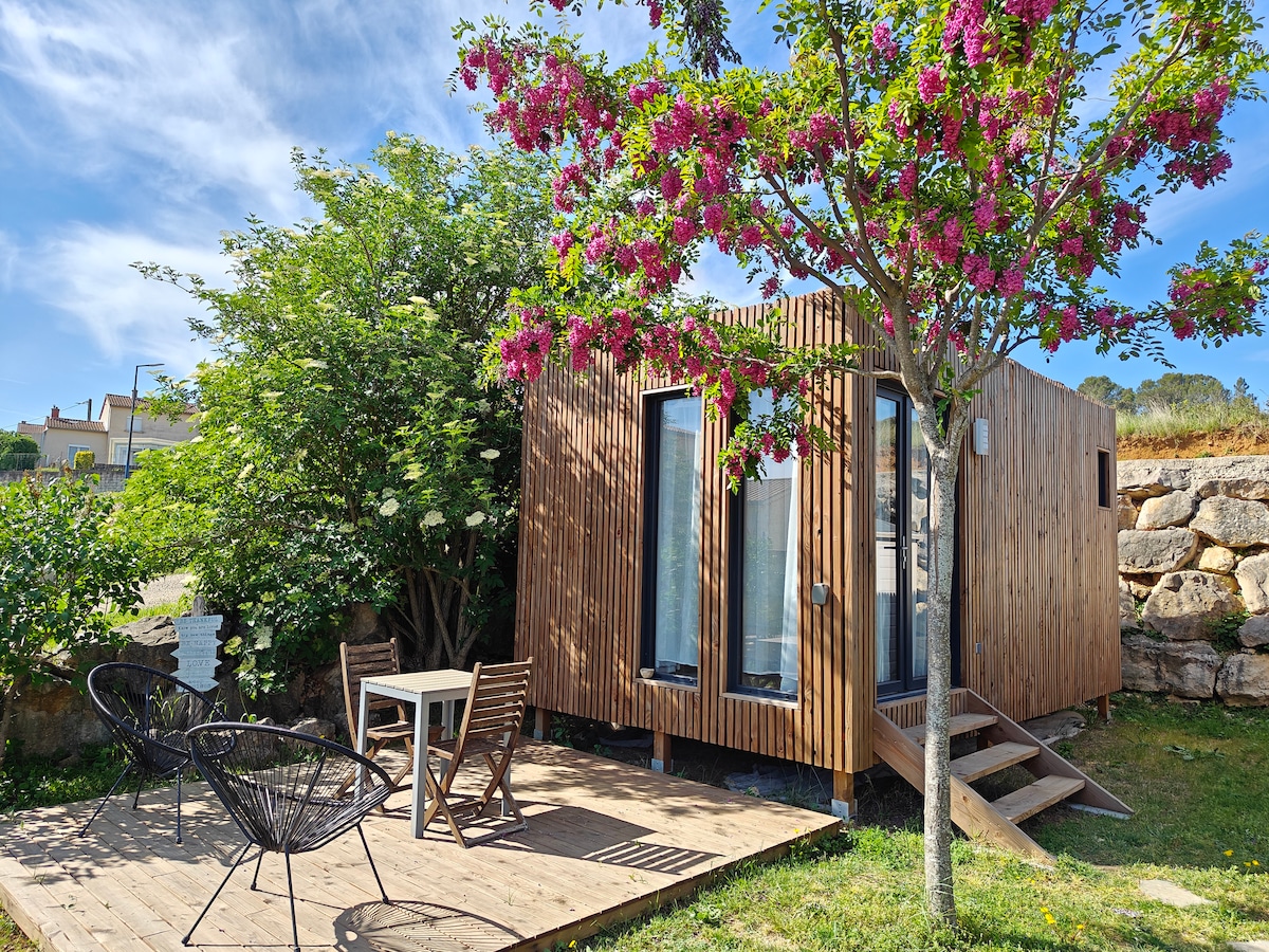 A modern wooden cabin is set against a lush backdrop, featuring large glass doors and a spacious wooden deck. A small outdoor table and four chairs are arranged under a blooming tree, adding a touch of colour to the serene environment.