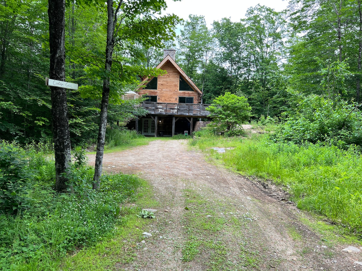 A spacious log cabin stands amidst a lush green landscape, framed by trees. A gravel path leads up to the entrance, showcasing a large wrap-around deck. Natural foliage surrounds the cabin, creating a peaceful and secluded environment.