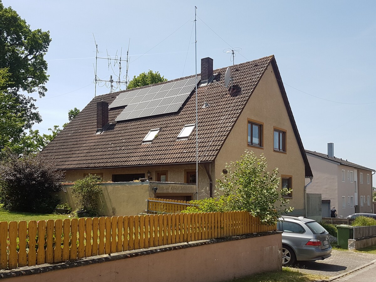 A two-story house is shown with a sloped roof featuring solar panels. Surrounding greenery enhances the outdoor area, while a wooden fence outlines the property. Nearby vehicles and neighboring structures are visible, providing context to the residential setting.