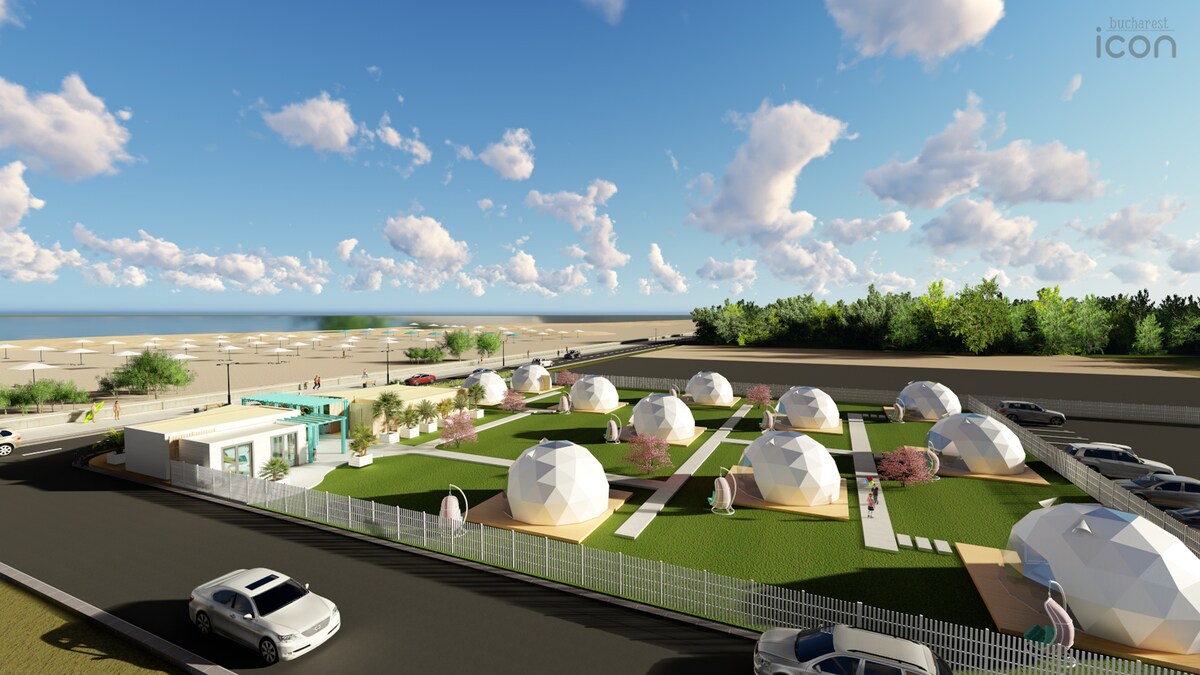 A wide view of a glamping site featuring multiple dome-shaped accommodations arranged on a grassy area. Nearby, a reception building is visible. The beach can be seen in the distance, with a clear sky and scattered clouds enhancing the serene outdoor setting.