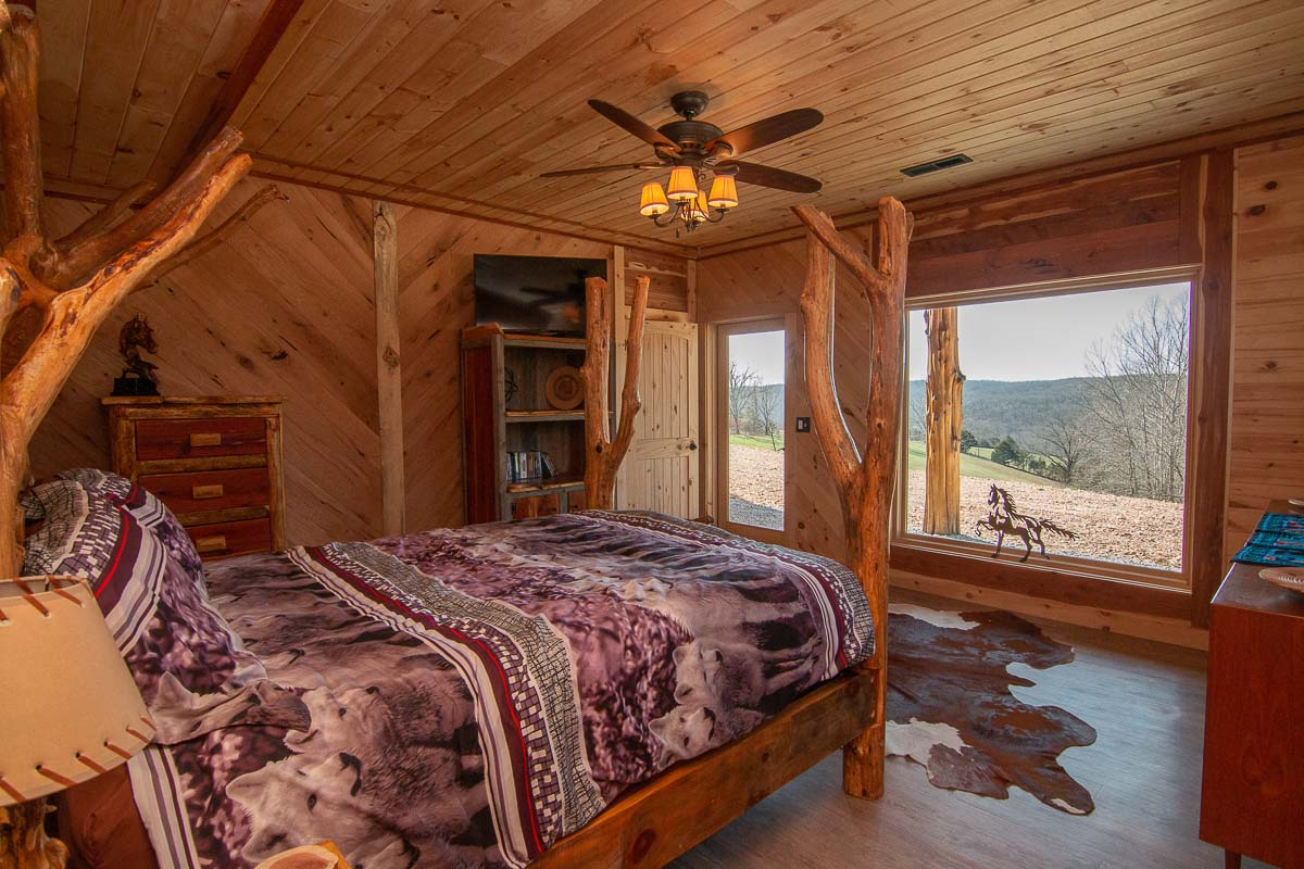 A rustic bedroom features log furniture, including a spacious bed adorned with a patterned blanket. A large window offers a scenic view of the surrounding landscape, while natural wood walls and ceiling create a warm ambiance. A TV and storage unit are present for convenience.
