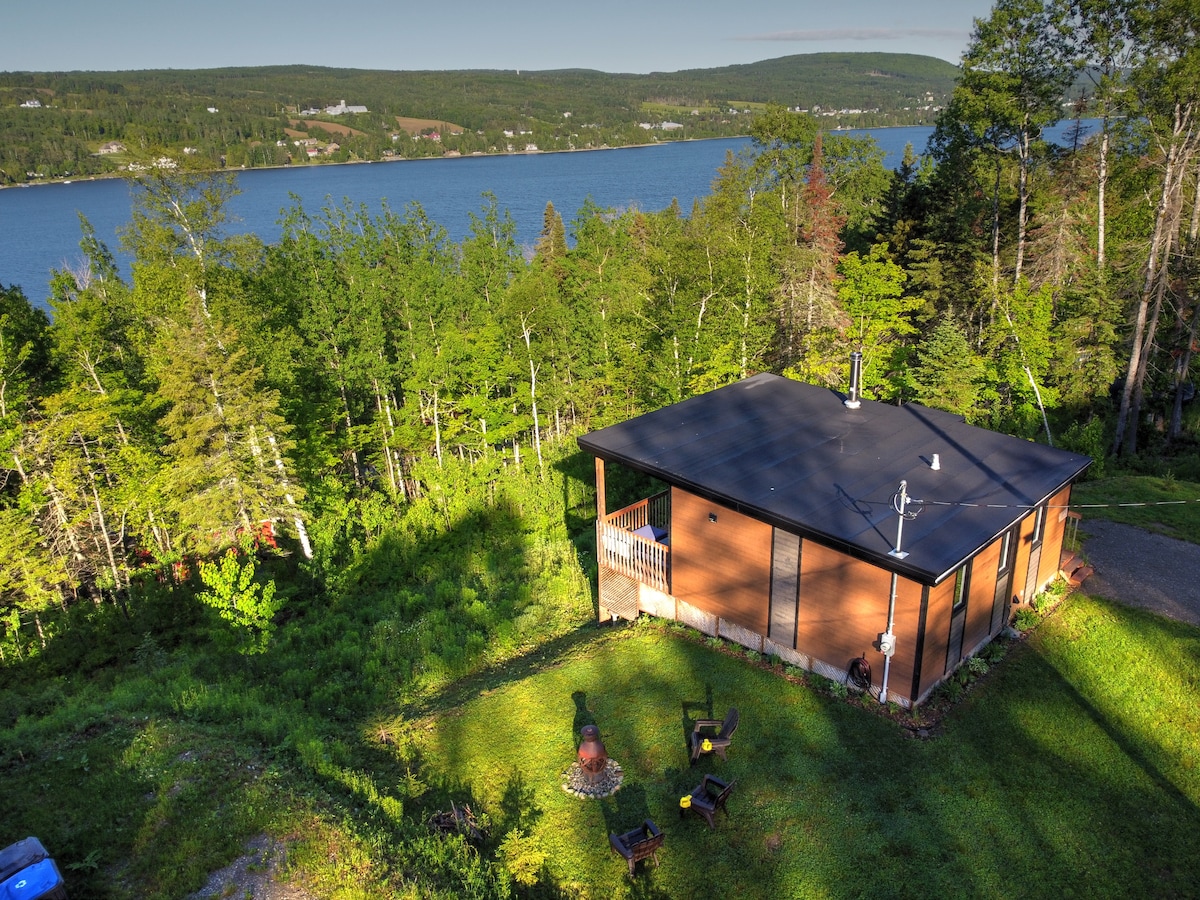 The cottage is set amidst lush greenery, overlooking Lake Pohénégamook. The wooden structure features a dark roof and a porch area. A fire pit and seating are visible in the foreground, enhancing the natural setting.