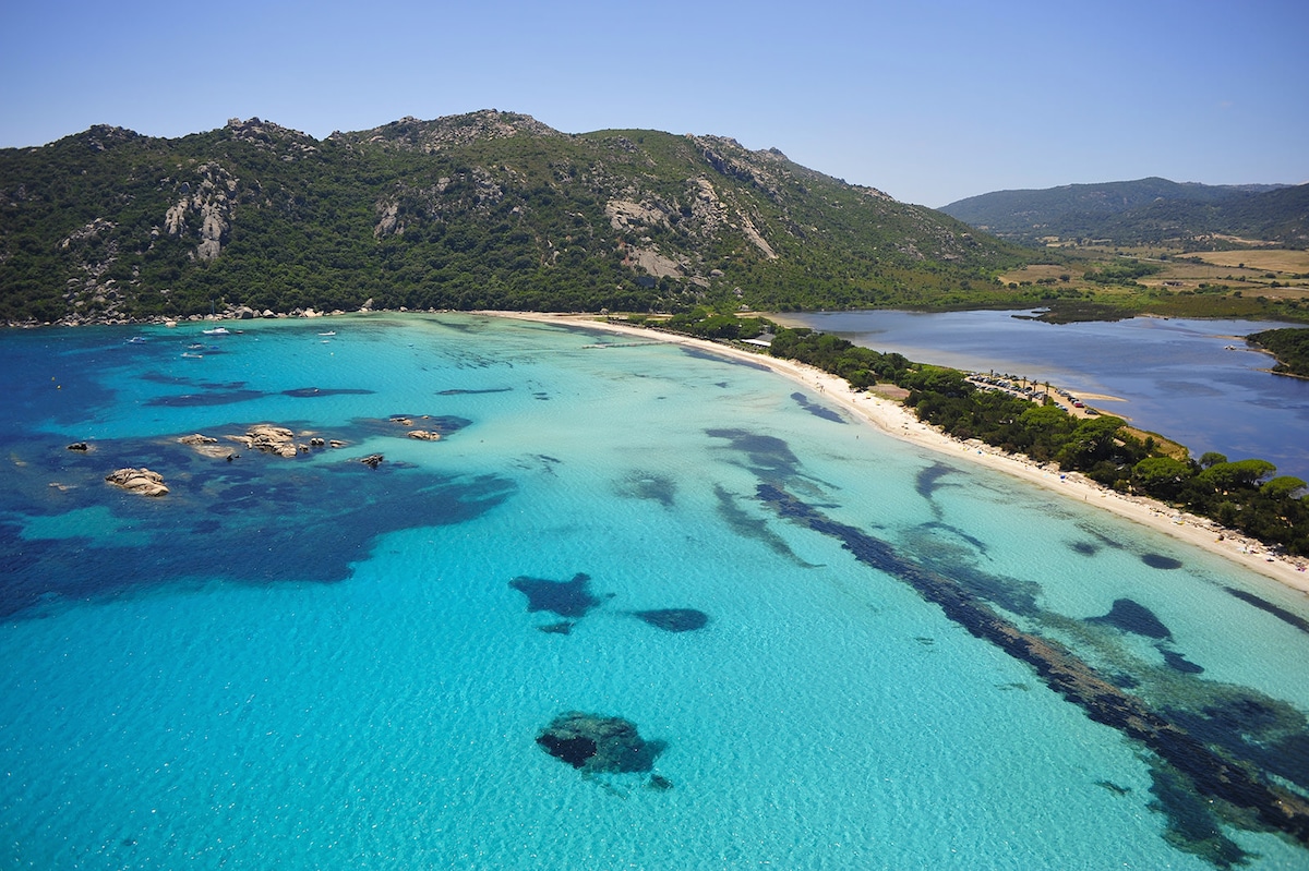 A serene beach view captures turquoise waters gently lapping against the shore, bordered by a lush green landscape and distant mountains. Rocky formations are visible in the water, adding texture to the shoreline. The scene showcases a combination of natural beauty and tranquility.