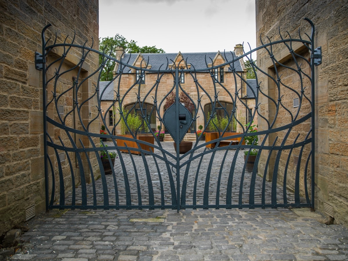 An ornate metal gate, designed with intricate leaf patterns, stands at the entrance of Ratho Park Steading. The cobblestone pathway is visible beyond the gate, leading into a charming courtyard framed by the stone structures of the property.