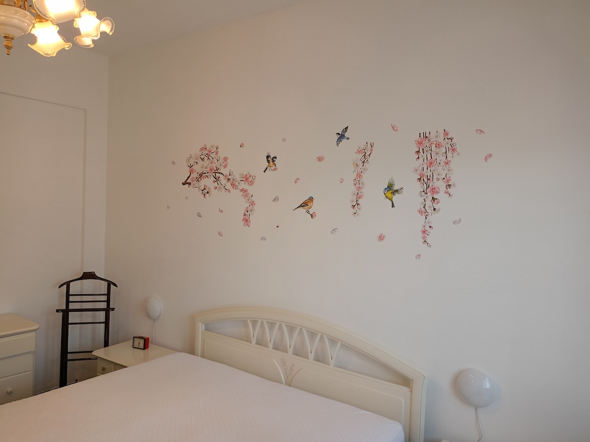 A well-lit bedroom features a spacious bed against a white wall adorned with delicate floral and bird decals. A bedside table with a lamp is present, along with a dresser and a wooden coat rack. Soft lighting from an overhead fixture creates a welcoming ambiance.