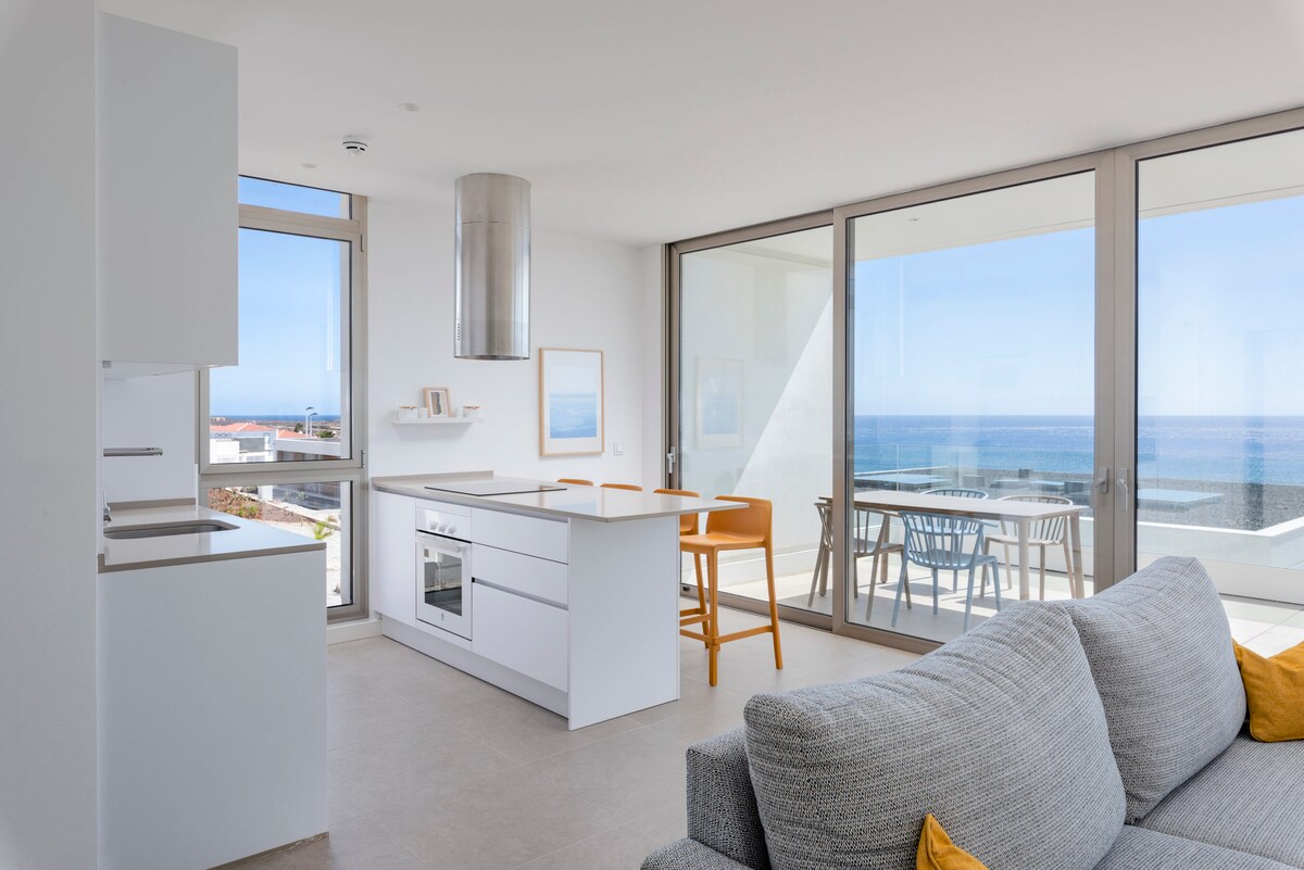 A modern living area is showcased, featuring a grey sofa and a minimalistic kitchen with a white cabinetry. Expansive glass doors provide a clear view of the ocean, with a dining area visible on the terrace, allowing natural light to fill the space.