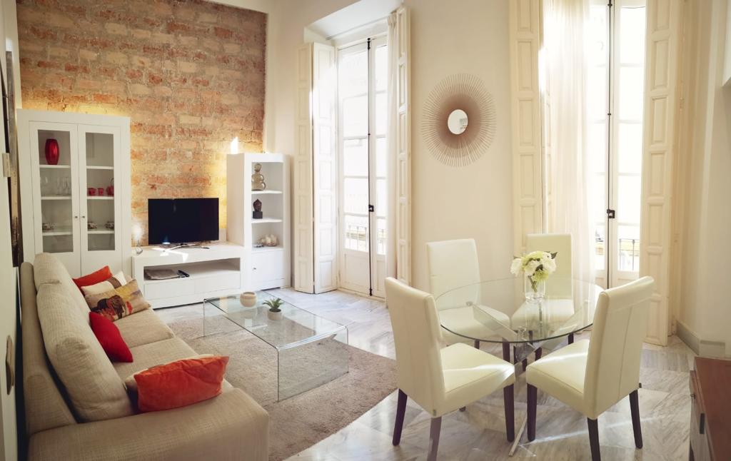 A spacious living area features a comfortable sofa with cushions, a glass coffee table, and a dining table surrounded by chairs. Natural light fills the room through large windows, while a textured brick wall adds visual interest. A flat-screen TV is positioned against a white cabinet.