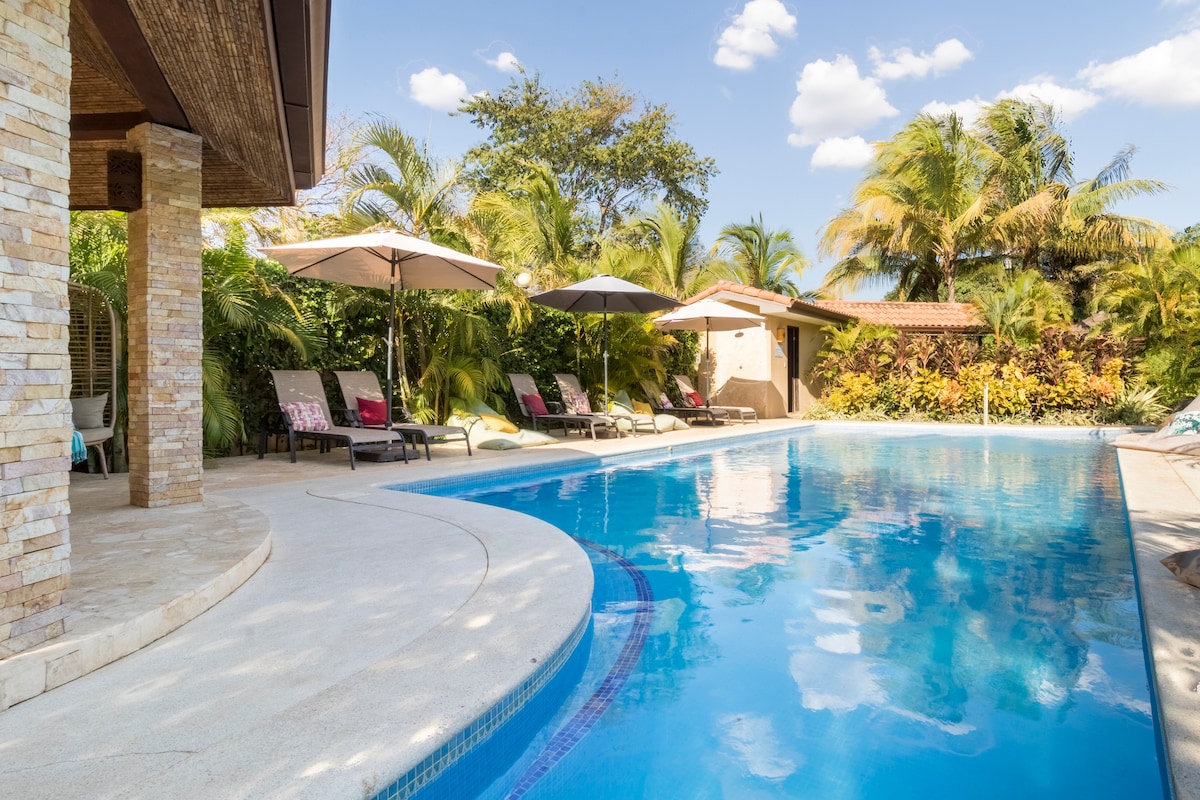 A serene outdoor space features a large swimming pool surrounded by tropical vegetation. Several lounge chairs under umbrellas are positioned along the pool’s edge, inviting relaxation. A sun-drenched patio area is visible, enhancing the tranquil setting.