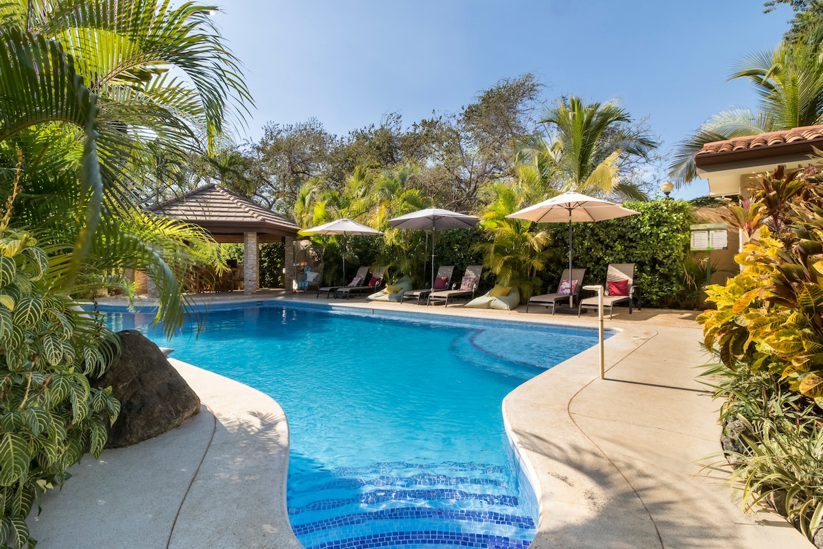 A serene pool area is surrounded by lush tropical foliage, featuring a clear blue pool with gentle curves. Two shaded seating areas with umbrellas are positioned beside the pool, offering comfortable lounge chairs for relaxation.