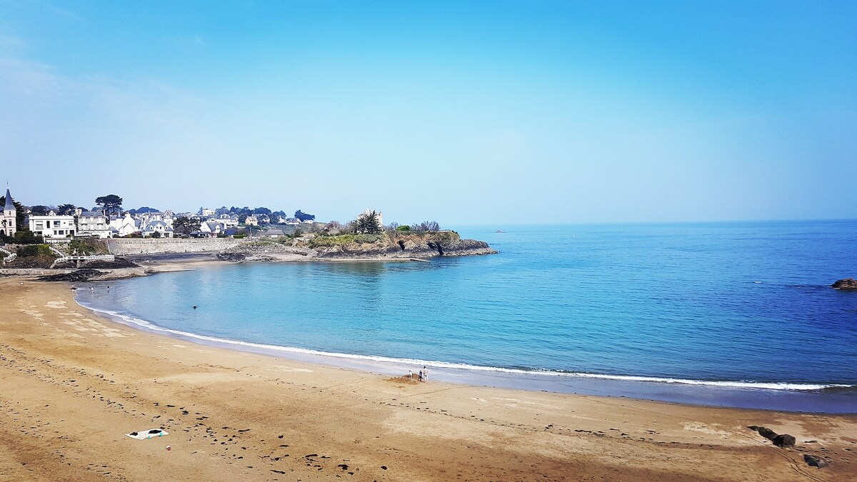 A serene beach scene is depicted with calm blue waters stretching towards the horizon, merging with a clear sky. Sandy shores gently curve along the water's edge, while a coastal area with scattered buildings and greenery provides a picturesque backdrop.