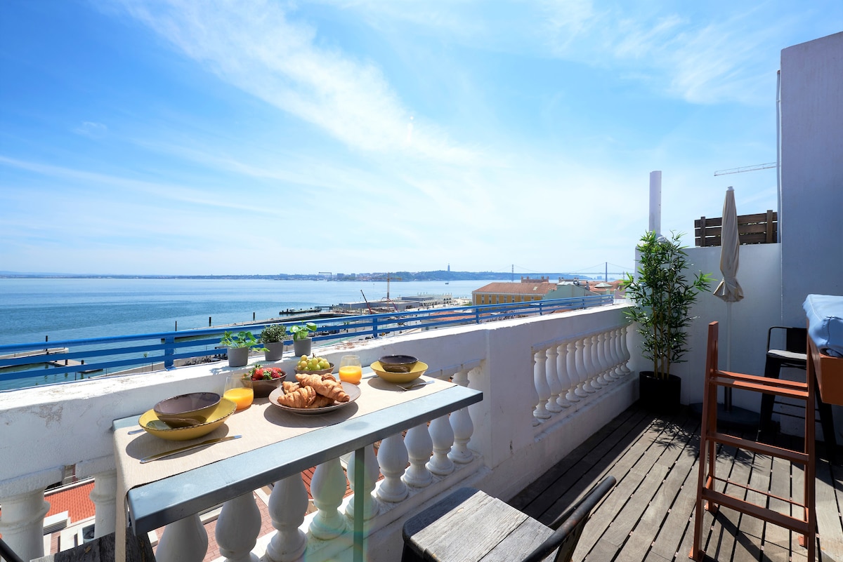 A rooftop terrace is displayed, featuring a dining table set for breakfast with plates, drinks, and pastries. The expansive river view captivates, framed by a clear blue sky, while stylish outdoor furniture complements the inviting space.
