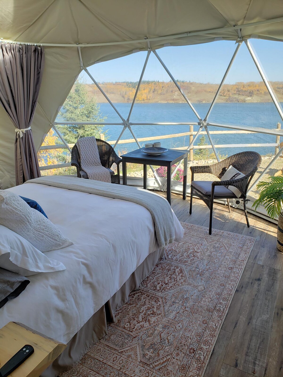 A geodesic dome interior features a spacious bed with soft linens, framed by large windows offering expansive views of the lake and surrounding landscape. A small table with two chairs is positioned nearby, complemented by a decorative rug and a potted plant for added warmth.