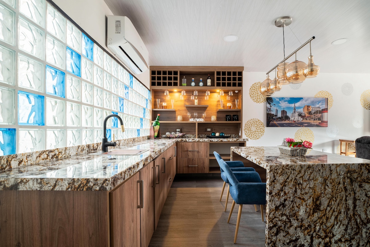 The modern kitchen features a stunning granite countertop with rich patterns, complemented by a glass-block wall allowing natural light. Two stylish blue chairs are set around a dining table, while shelves display decorative items and glassware, creating an inviting cooking and dining space.