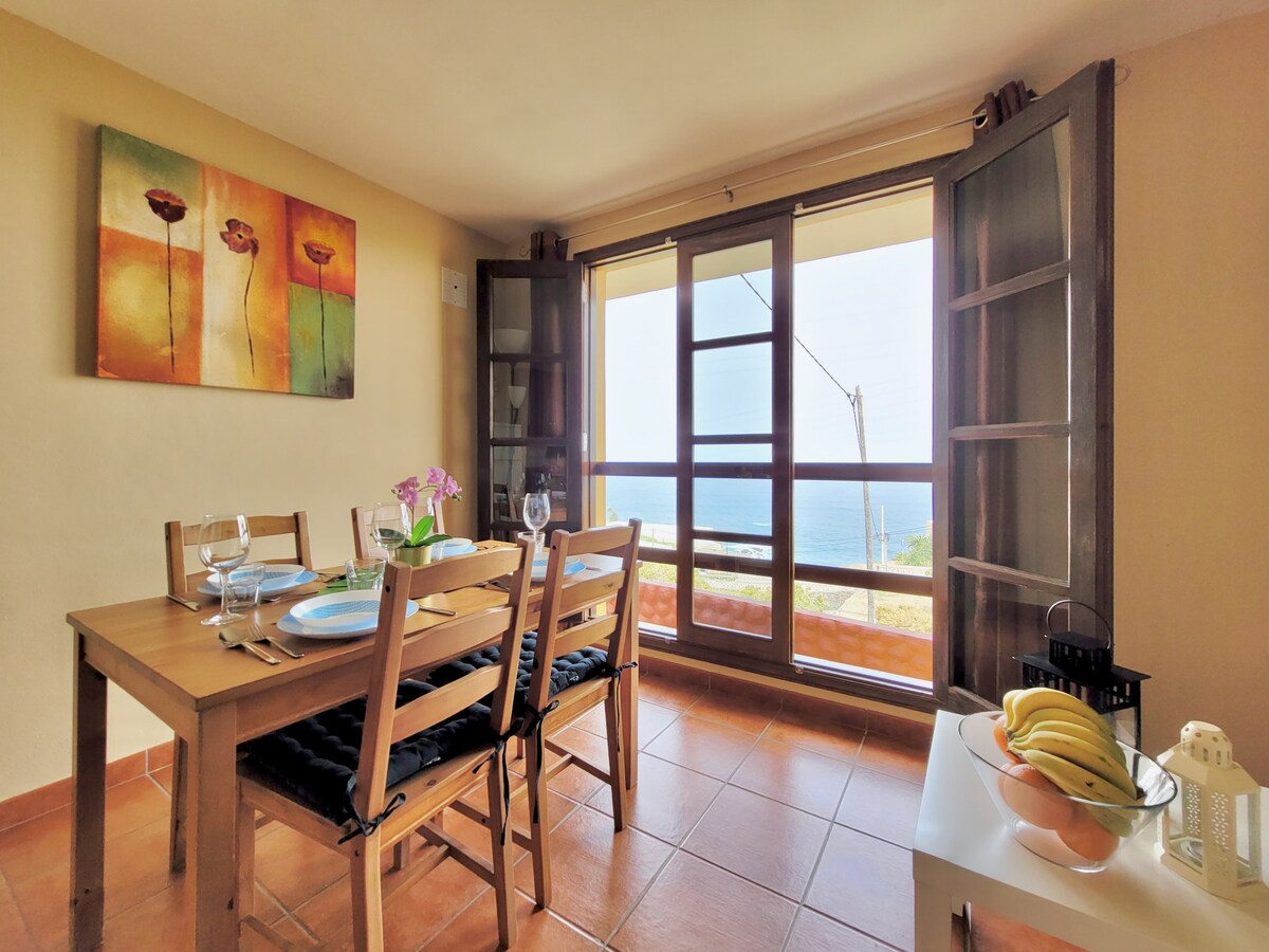 A dining area features a wooden table set for four, with plates and glasses placed on a neatly arranged tablecloth. Large wooden windows provide a view of the sea, while a floral painting adds color to the wall. A bowl of bananas and decorative lantern are visible in the room.