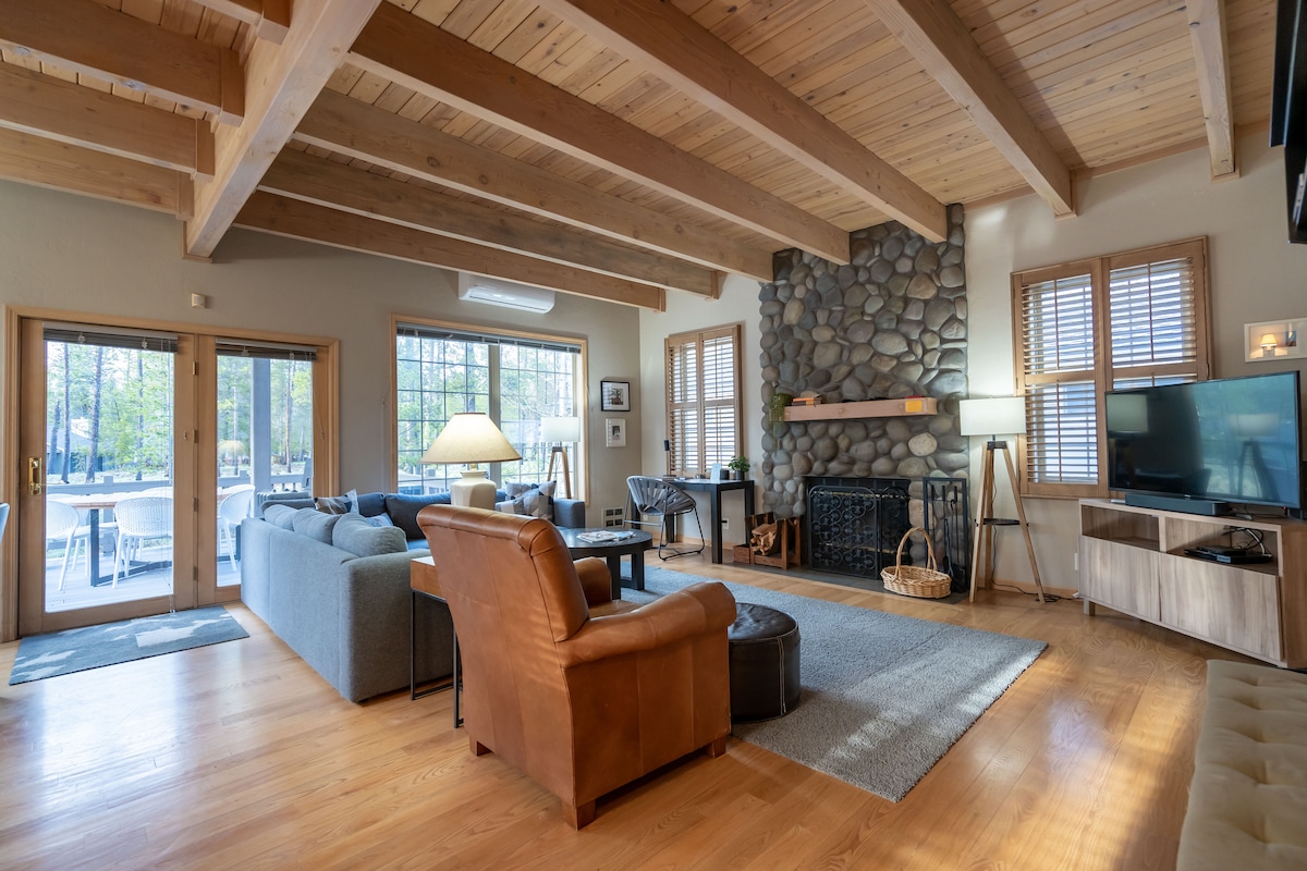 The living area features high tongue and groove wood ceilings supported by exposed beams. A cozy wood burning fireplace made of river rock serves as a focal point, with a large window offering views of the natural surroundings. Comfortable seating arrangements are present, including a leather armchair and a sofa.
