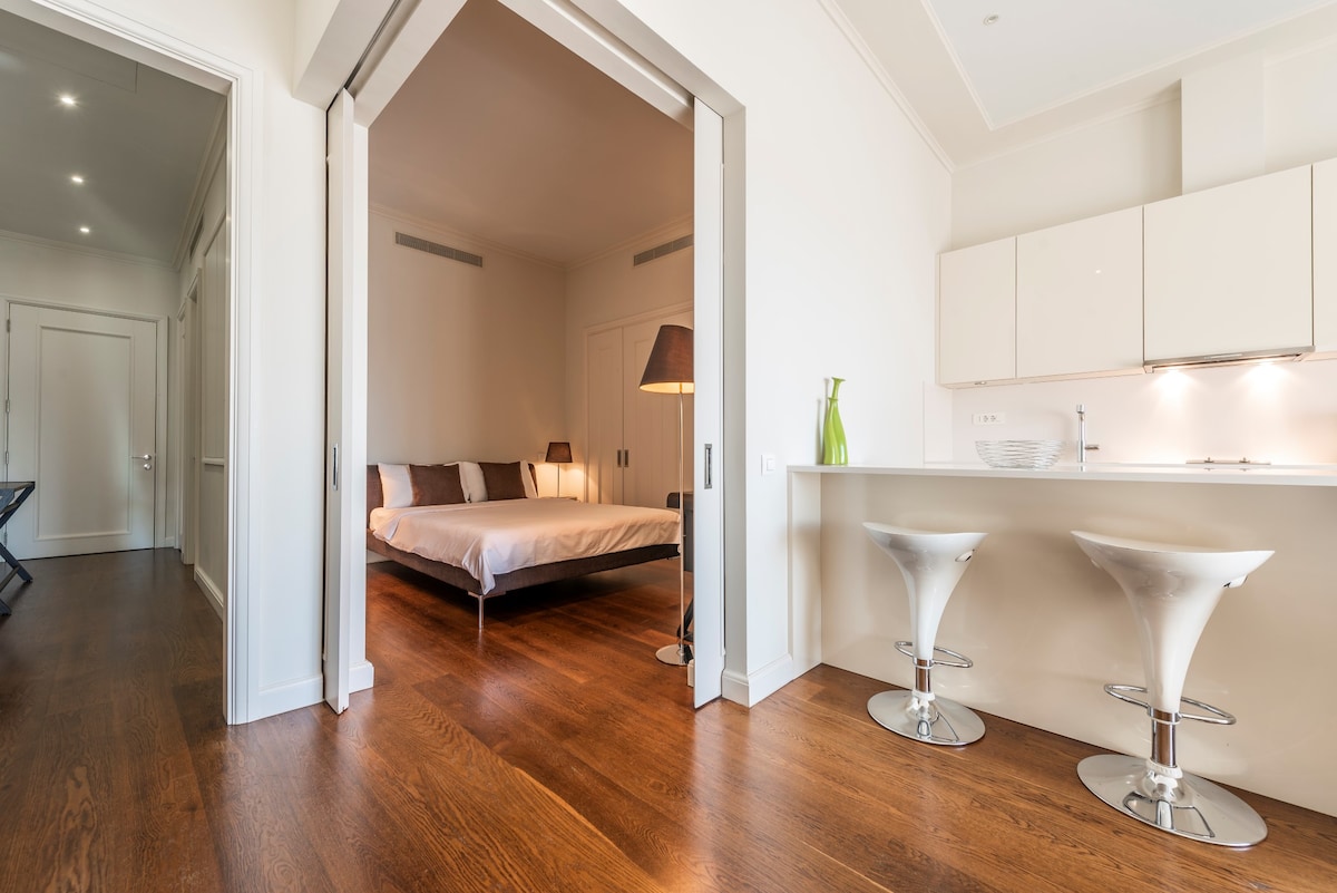 A spacious living area features a modern kitchen island with two stylish bar stools. Soft natural light fills the room, highlighting a cozy bedroom with a large bed positioned against the wall. Light wood flooring contrasts with the bright walls and minimalist decor.