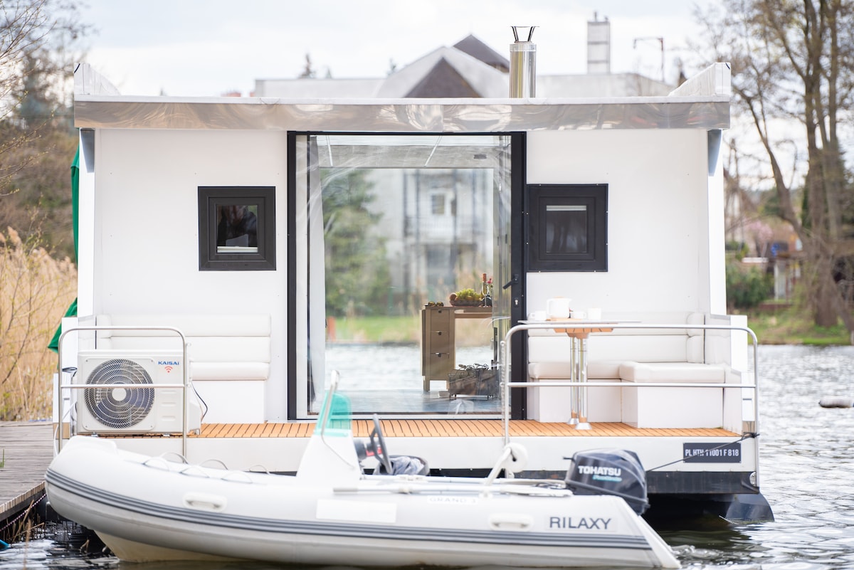A modern houseboat is anchored in a serene setting, featuring large glass doors that open to a deck with comfortable seating. A small table is positioned next to the seating area, allowing for outdoor dining. A motorboat is docked alongside, enhancing accessibility.