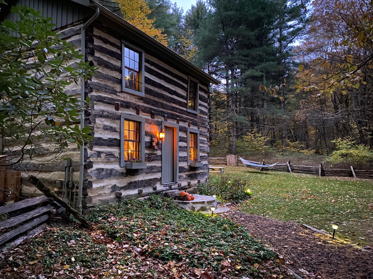 The historic log cabin is nestled among trees, showcasing its charming wooden exterior and multiple small windows. Soft lighting illuminates the entrance, while a hammock and a grassy yard invite relaxation in the serene outdoor space.