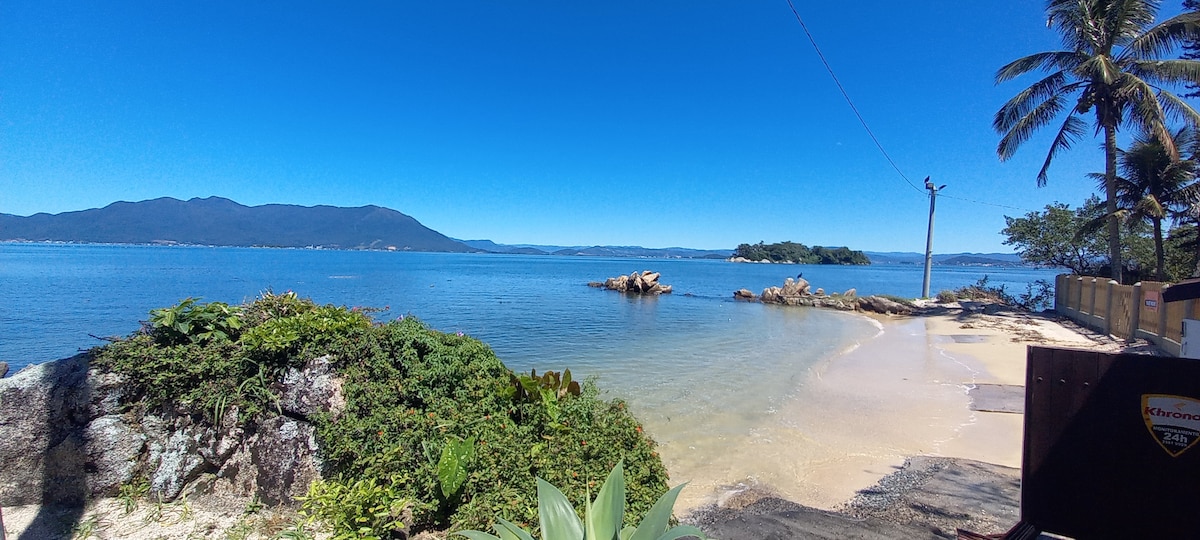 A serene beach scene is depicted, featuring calm, clear waters that gently meet a sandy shore. Lush greenery frames the view, with distant hills visible under a clear blue sky. A rocky outcrop is seen offshore, adding to the tranquil coastal landscape.
