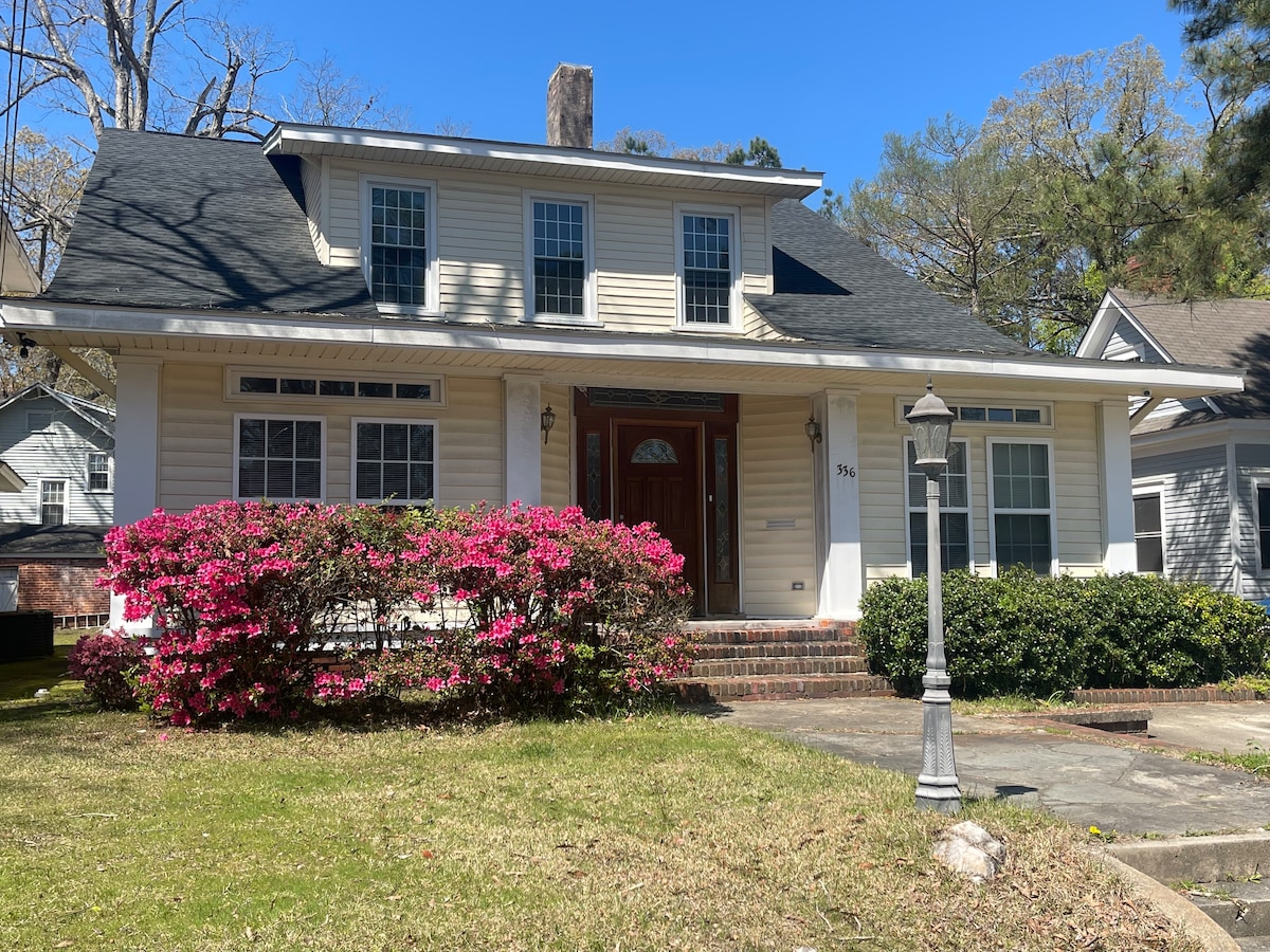 The exterior of a two-story home is showcased, featuring white siding and a welcoming front porch. Bright pink azaleas bloom in the garden, adding color to the landscaping, while a decorative lamp post stands along the pathway leading to the entrance.