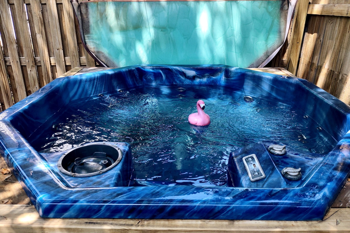 A private hot tub sits within a wooden enclosure, featuring a vibrant blue interior. A playful pink flamingo floats on the water's surface, accompanied by built-in controls beside the tub for guest convenience.