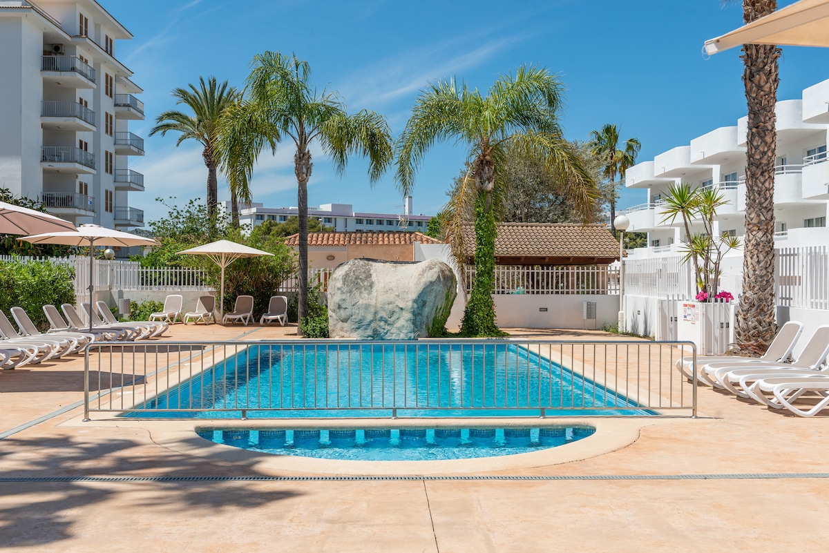 A well-maintained private pool is surrounded by palm trees and lounge chairs. Sun umbrellas provide shaded areas near the water. A rock feature adds a natural element to the setting. The pool area is bright and open, with clear blue water inviting relaxation.