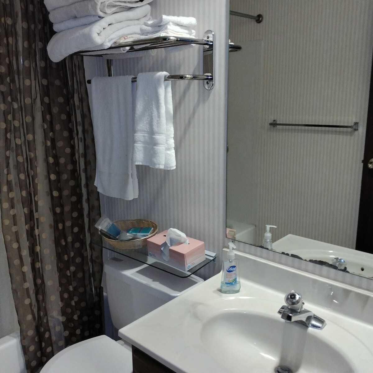 A well-appointed bathroom features a white sink with a chrome faucet. Fresh towels are neatly arranged on a chrome rack, with additional toiletries placed on a shelf nearby. A bathtub is partially visible, accompanied by a shower curtain with a polka dot pattern.