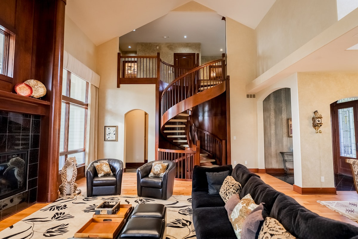 The spacious living area features a grand staircase made of rich wood, leading to an upper level. A dark sofa is arranged around a coffee table, complemented by two armchairs. Large windows allow natural light to fill the room, illuminating decorative elements within.