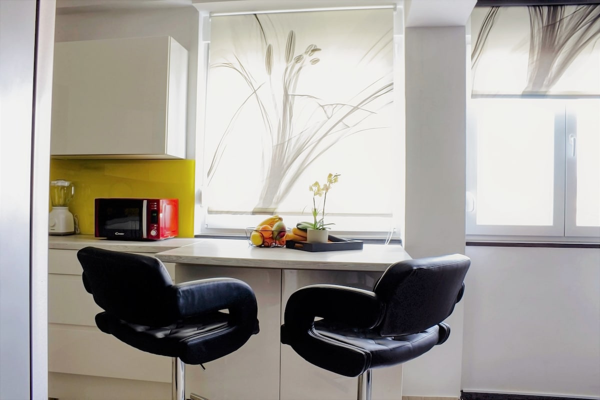 A modern kitchen space features a small bar table with two black swivel chairs. Light-colored cabinetry contrasts with a vibrant yellow accent wall. Natural light pours in through two windows adorned with floral-patterned shades, showcasing fresh fruit on the table.