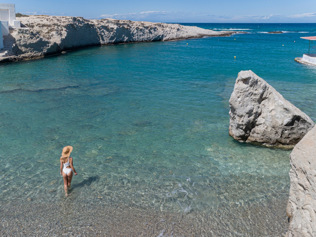 A calm, clear water shoreline is depicted, featuring smooth stones and rocky formations. A person is seen wading into the refreshing water, with gentle waves lapping at the shore. Lush blue sea extends into the horizon, under a bright, clear sky.