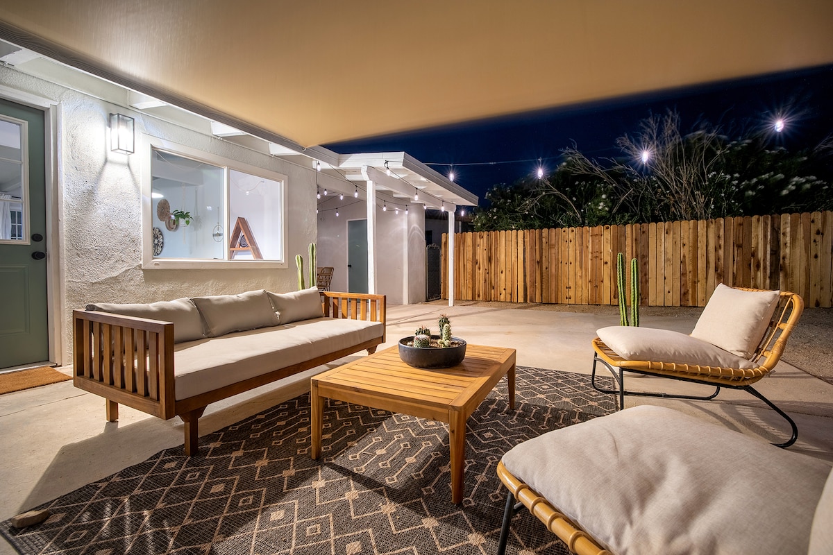 An outdoor lounge area is illuminated by soft lighting, featuring a wooden coffee table surrounded by a spacious sofa and a single lounge chair. A textured area rug anchors the seating arrangement, while a wooden fence and potted cacti frame the space.