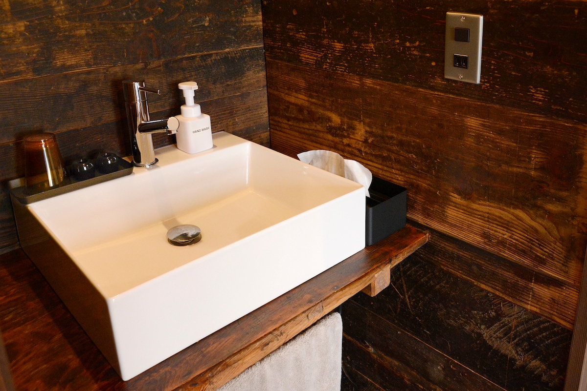 A modern sink is situated atop a wooden countertop in a bathroom with rustic wooden walls. A soap dispenser and a tissue box are positioned nearby, complemented by a small wastebasket. The space offers a functional, clean appearance.
