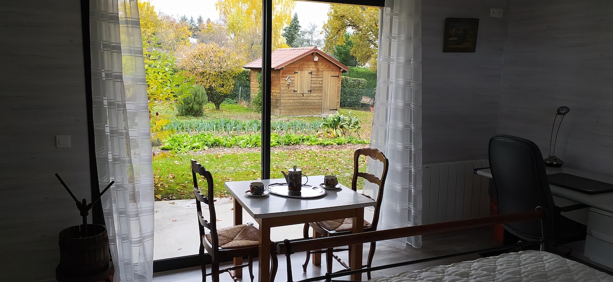 A spacious room is highlighted by large glass doors that open to a garden view, showcasing autumn foliage. A small table set for two is positioned near the doors, with wooden chairs and a desk area visible in the interior.