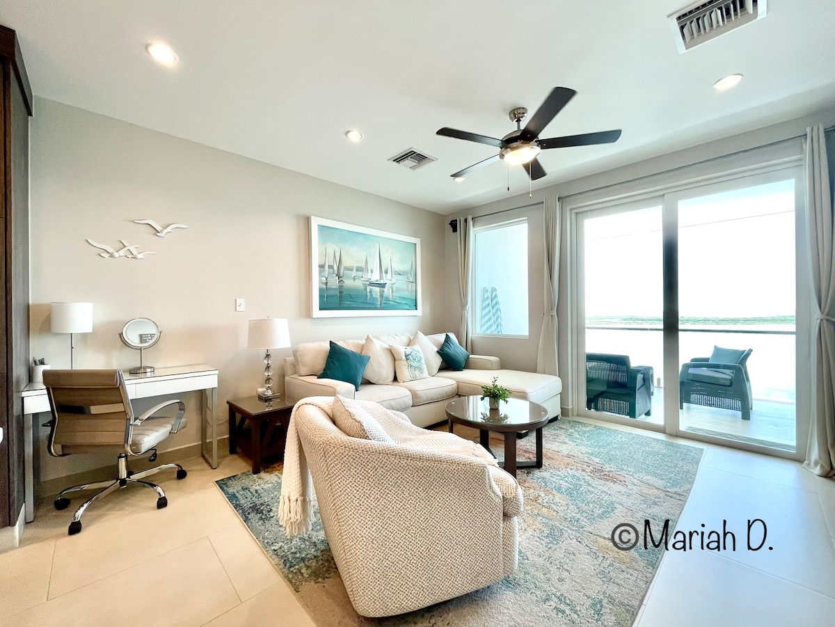 A spacious living area features a comfortable sofa surrounded by soft cushions. A desk with a chair is positioned next to a bright window, while light curtains enhance the natural light. Artistic decorations and a patterned rug add a touch of elegance to the room.