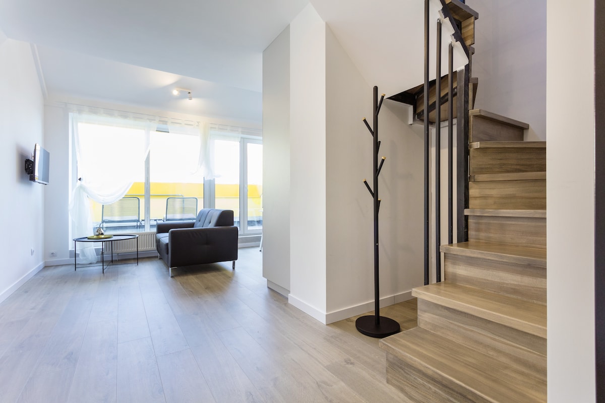 A modern interior showcases a spacious living area with large windows allowing natural light to fill the room. A comfortable sofa is positioned near the window, while a sleek staircase leads to the upper floor. Minimalist decor includes a coat rack placed beside the stairs.