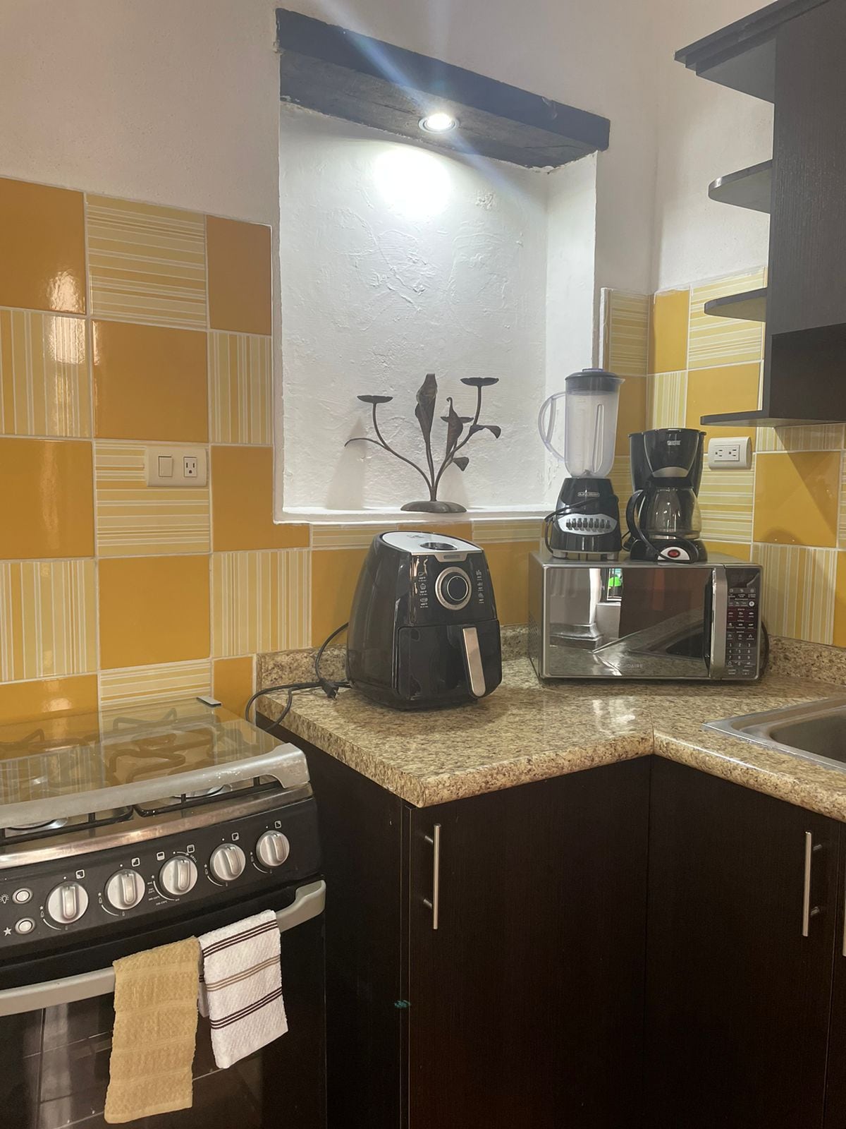 A compact kitchen area features modern appliances, including a stove, microwave, blender, and coffee maker. Brown cabinets complement the yellow and white patterned backsplash. A sink is integrated into the granite counter, providing an efficient workspace.