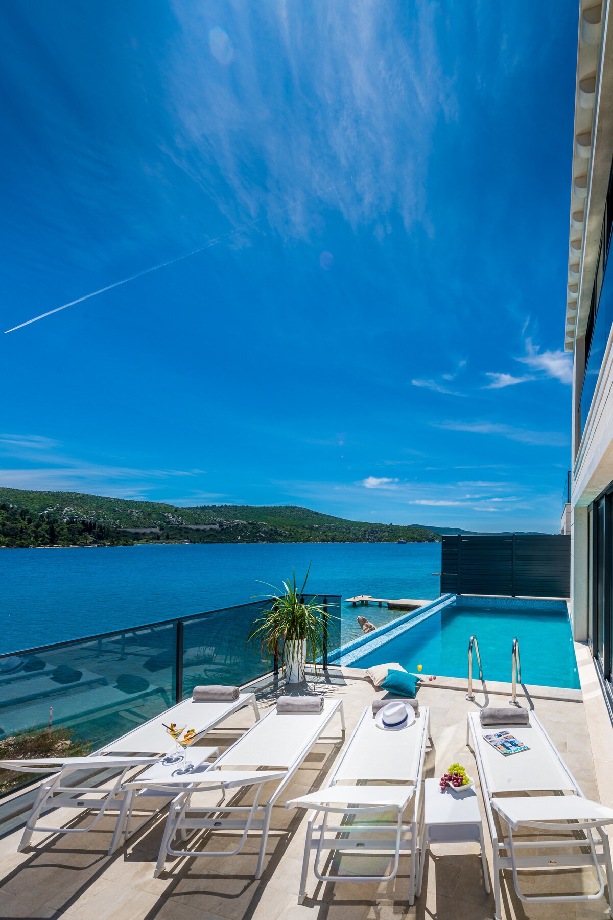 An outdoor terrace features several sun loungers positioned near an infinity pool overlooking the blue Adriatic Sea. The space is surrounded by glass railings, with vibrant greenery adding a touch of nature. The clear blue sky enhances the feeling of relaxation.