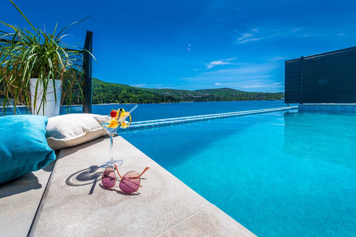 An infinity pool reflects the clear blue sky, with green hills visible beyond the water's edge. A glass of fruit cocktail rests on the poolside next to a pair of sunglasses and a soft, blue pillow, inviting relaxation in the serene atmosphere.
