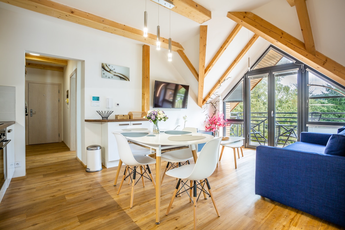 A spacious living area is illuminated by natural light streaming through large glass doors, which lead to a balcony. A modern dining table is surrounded by white chairs, while a blue sofa complements the warm wooden accents of the exposed beams and flooring.